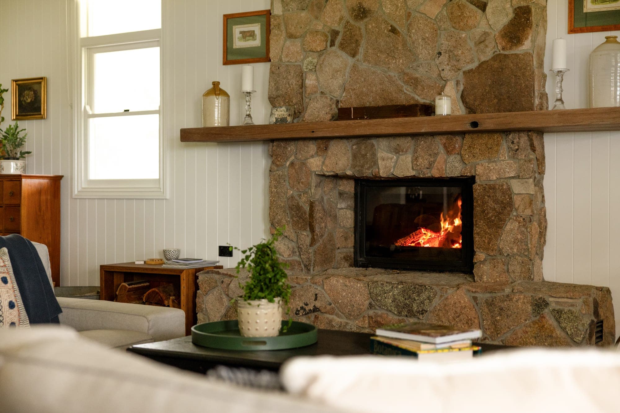 Living room stone feature fireplace with glowing fire, timber mantel beam and white vertical board walls
